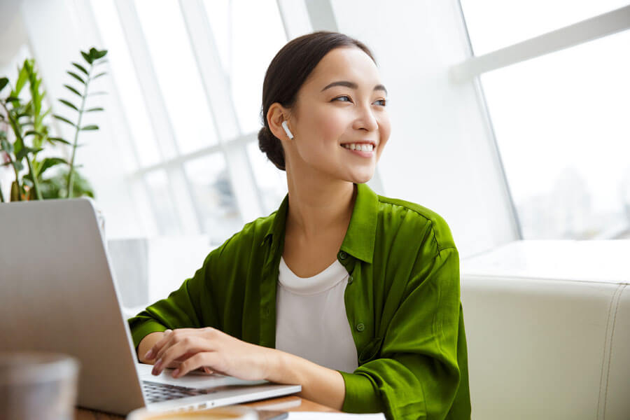 Woman with earpods smiling and typing on laptop in bright room